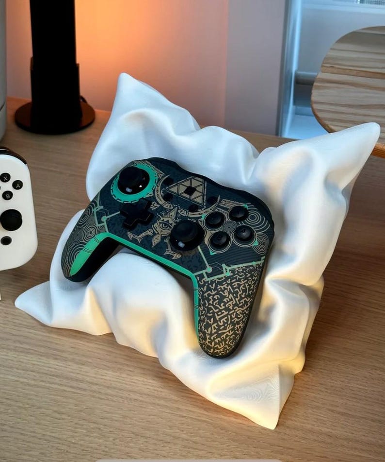 May include: A black and green video game controller with a decorative design featuring a green triangle and gold symbols. The controller is resting on a white pillow.