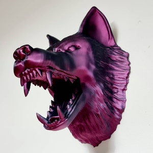 May include: A 3D-printed wolf head sculpture in shades of purple and black, mounted on a wall. The wolf's mouth is open, revealing sharp teeth. The sculpture is a decorative item.