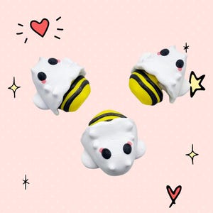 May include: Three white and yellow bee figurines with black eyes and pink cheeks. The bees are in a curled up position, as if they are sleeping.