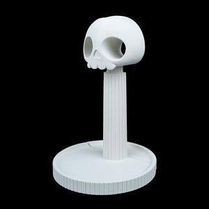 May include: A white, decorative skull-shaped object with large eye sockets and a detailed jawline. The skull sits atop a vertical, ridged post, which is connected to a circular base with a similar ridged design. The object is set against a black background.