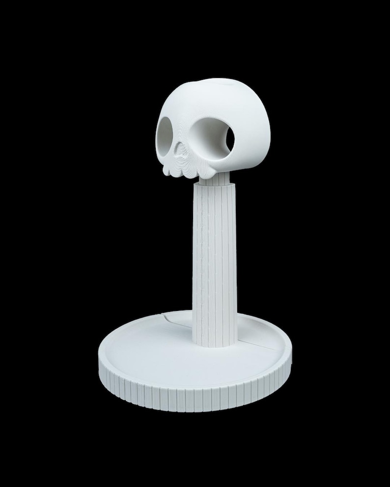 May include: A white, decorative skull-shaped object with large eye sockets and a detailed jawline. The skull sits atop a vertical, ridged post, which is connected to a circular base with a similar ridged design. The object is set against a black background.