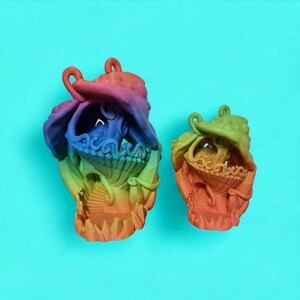 May include: Two colorful ceramic sculptures of fantastical creatures with intricate details. The larger sculpture is a vibrant rainbow of colors, while the smaller sculpture is a more muted rainbow with a green hue. Both sculptures have a whimsical and organic design.