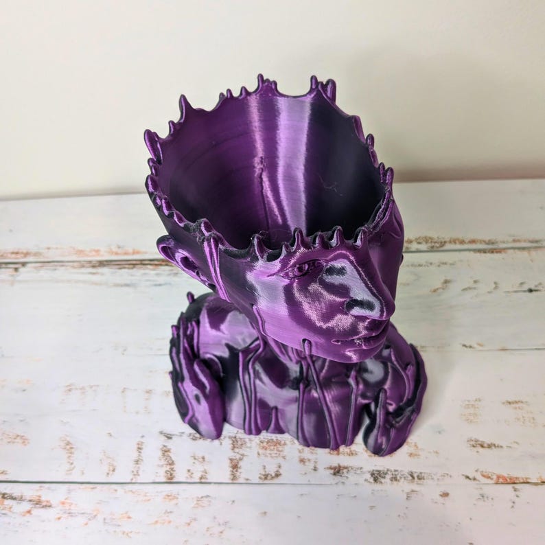 May include: A 3D-printed, purple planter shaped like a human head. The planter has a detailed face and a textured surface. The top of the head forms a wide opening for plants. The design includes a crown-like edge.