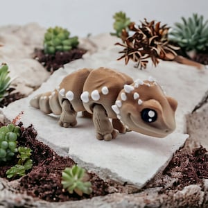 May include: A small, brown and white toy lizard with a segmented body and large, black eyes. The lizard is posed on a light gray rock surface.