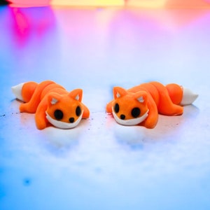 May include: Two orange and white fox figurines lying on a blue surface. The foxes have black eyes and white noses.
