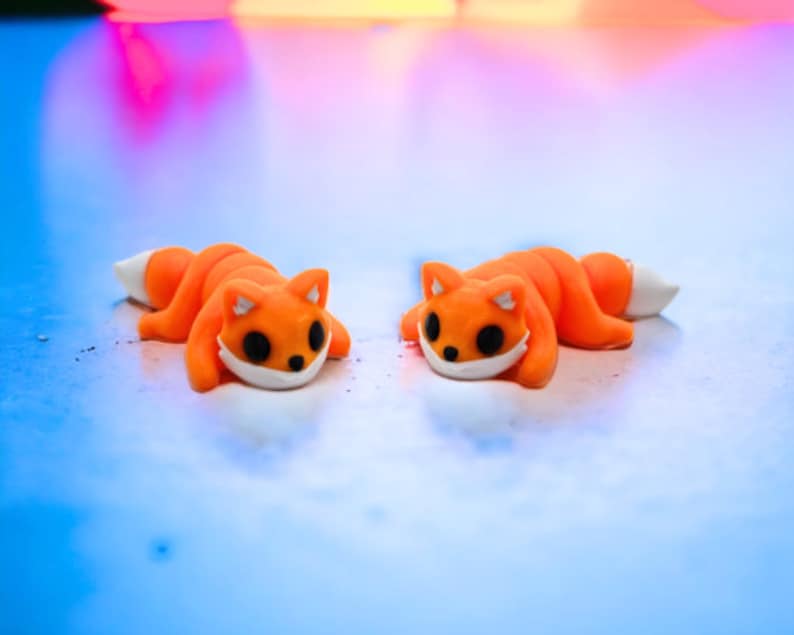 May include: Two orange and white fox figurines lying on a blue surface. The foxes have black eyes and white noses.