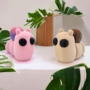 May include: Two small, 3D printed, plush toys shaped like snails. The snail on the left is pink with a purple and pink shell. The snail on the right is beige with a brown shell.