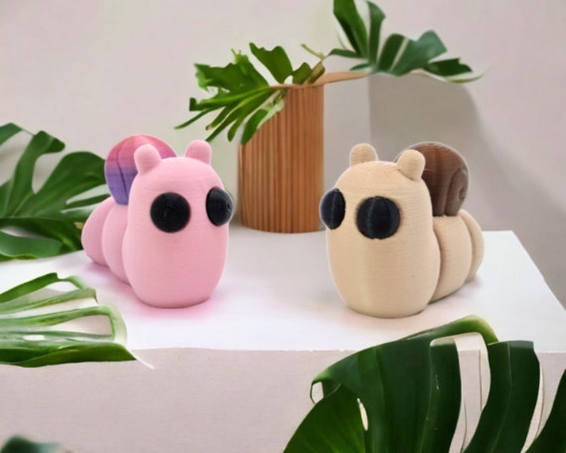 May include: Two small, 3D printed, plush toys shaped like snails. The snail on the left is pink with a purple and pink shell. The snail on the right is beige with a brown shell.
