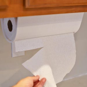 May include: A white paper towel roll mounted under a wooden cabinet. A hand is reaching for a sheet of the paper towels. The paper towels are white and have a textured surface.