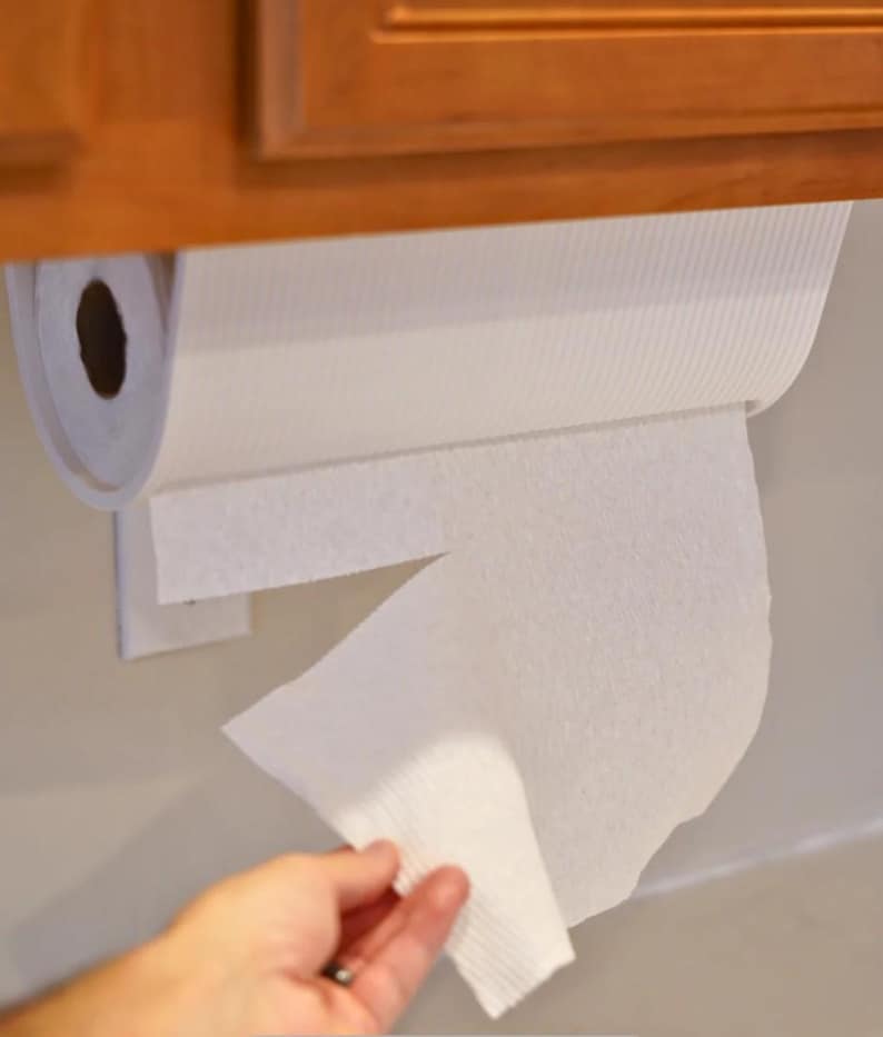 May include: A white paper towel roll mounted under a wooden cabinet. A hand is reaching for a sheet of the paper towels. The paper towels are white and have a textured surface.