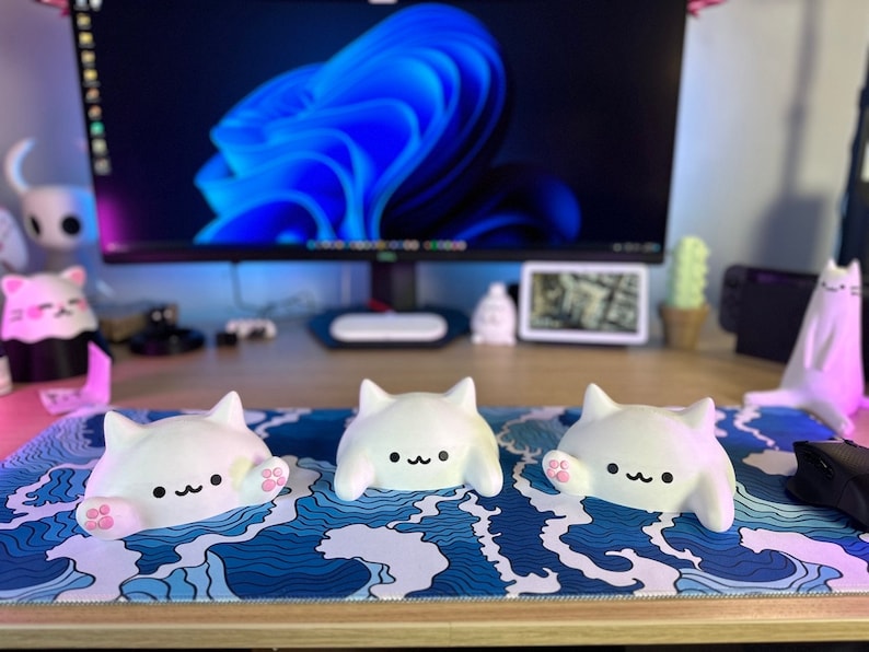 May include: Three white cat-shaped figurines with black facial features and pink paws are arranged on a blue and white wave-patterned desk mat. A computer monitor and other decorative items are in the background.