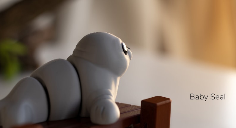 May include: A gray baby seal figurine with black eyes, sitting on a wooden surface. The figurine is made of a soft, pliable material and has a realistic appearance.  Baby Seal.