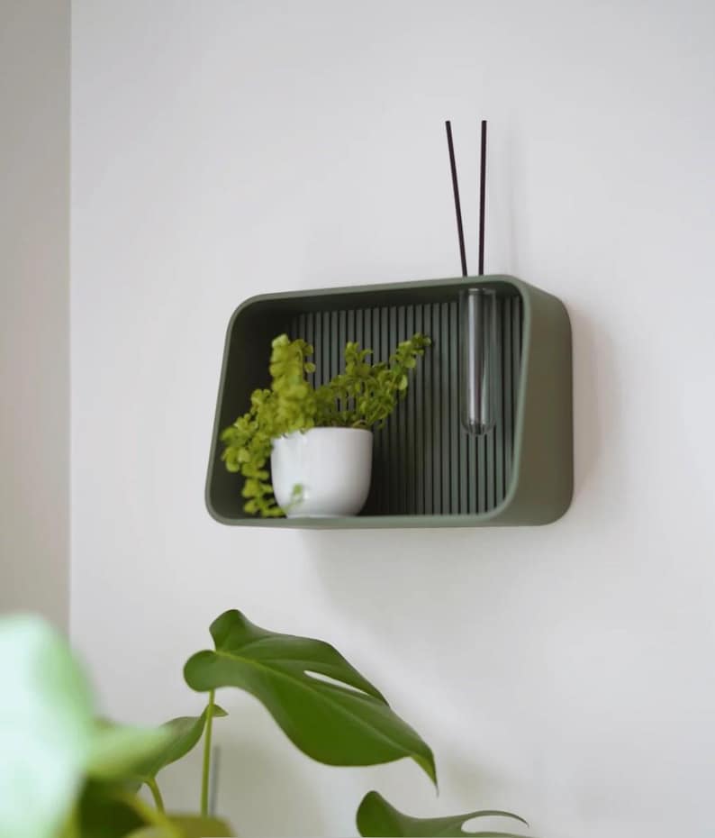 May include: A modern, olive green wall shelf with a built-in vase and a small white pot holding a green plant. The shelf has a vertical slatted back and is mounted on a white wall. Two black sticks are in the vase.
