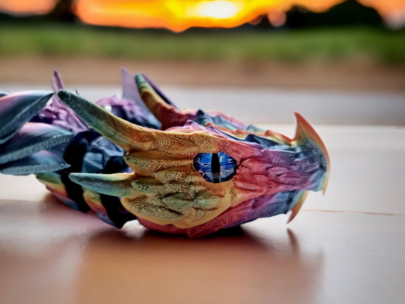 May include: A colourful dragon head figurine with a blue eye, made using a 3D printer. The dragon head is made of a rainbow of colours, including pink, yellow, blue, and green.