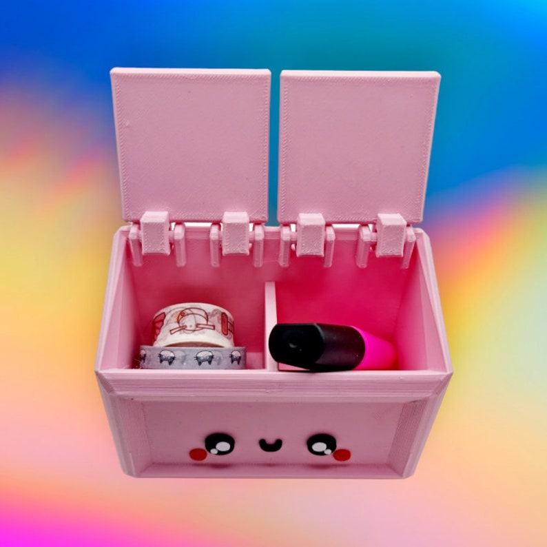 May include: A pink plastic box with a hinged lid, divided into two compartments. The box has a cute cartoon face on the front and contains a roll of washi tape and a pink highlighter.
