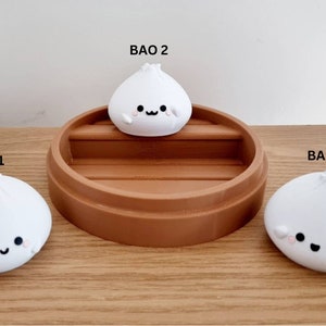 May include: Three white ceramic bao buns with cute faces, sitting on a brown wooden surface. The middle bao bun is on top of a brown wooden steamer.