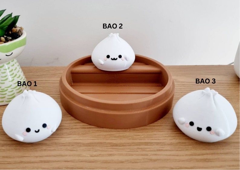 May include: Three white ceramic bao buns with cute faces, sitting on a brown wooden surface. The middle bao bun is on top of a brown wooden steamer.