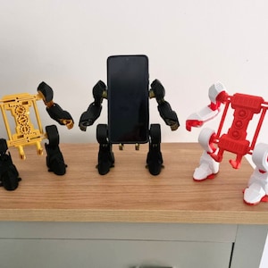 May include: Three 3D printed robot phone holders, one in black, one in gold, and one in white and red. The robots are holding a black smartphone in the middle.