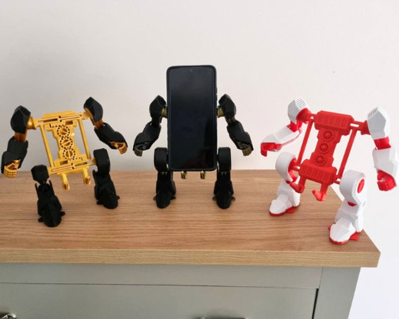 May include: Three 3D printed robot phone holders, one in black, one in gold, and one in white and red. The robots are holding a black smartphone in the middle.