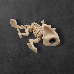 May include: A beige chameleon skeleton figurine with a blue and green eye. The figurine is posed in a walking position with its tail curled.