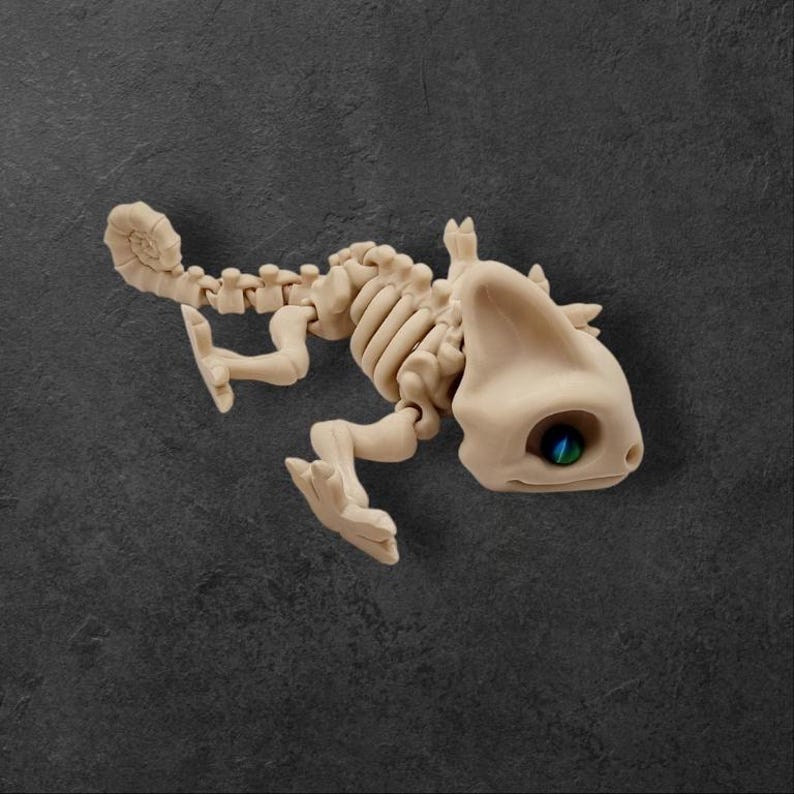 May include: A beige chameleon skeleton figurine with a blue and green eye. The figurine is posed in a walking position with its tail curled.