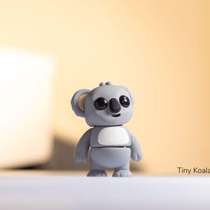 May include: A small gray koala figurine with black eyes and a white belly. The figurine is standing on a white surface. The text "Tiny Koala" is visible in the lower right corner of the image.