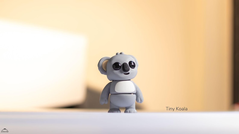 May include: A small gray koala figurine with black eyes and a white belly. The figurine is standing on a white surface. The text "Tiny Koala" is visible in the lower right corner of the image.