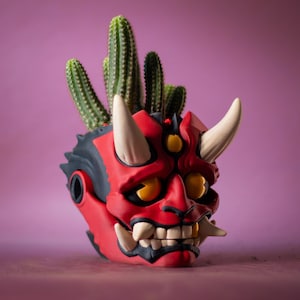 May include: A red and black Oni mask-shaped planter with white horns and teeth, holding several green cacti. The planter has yellow eyes and is set against a purple background.
