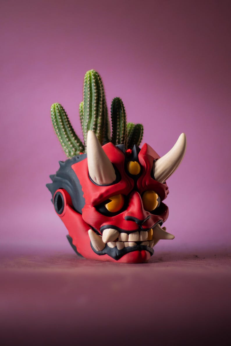May include: A red and black Oni mask-shaped planter with white horns and teeth, holding several green cacti. The planter has yellow eyes and is set against a purple background.