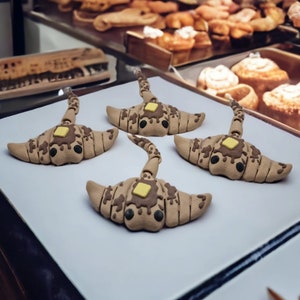 Op de afbeelding: Vier grillige croissantvormige sculpturen met bruine en beige details, bekroond met een vierkant stukje gele boter en chocoladedruppel. De sculpturen worden gepresenteerd op een wit oppervlak, met gebak op de achtergrond.