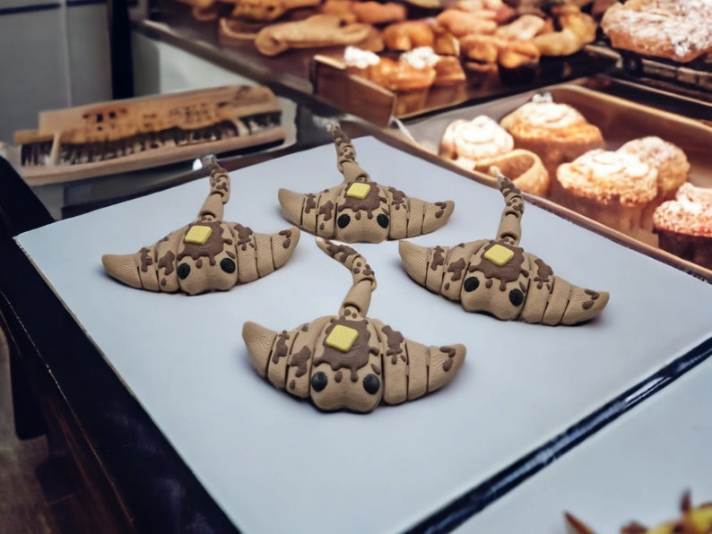 Op de afbeelding: Vier grillige croissantvormige sculpturen met bruine en beige details, bekroond met een vierkant stukje gele boter en chocoladedruppel. De sculpturen worden gepresenteerd op een wit oppervlak, met gebak op de achtergrond.