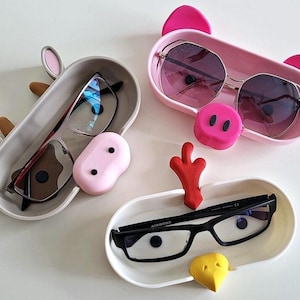 May include: Three colorful animal-shaped eyeglass holders. A pink pig, a brown cow, and a white chicken. Each holder has a pair of glasses inside.