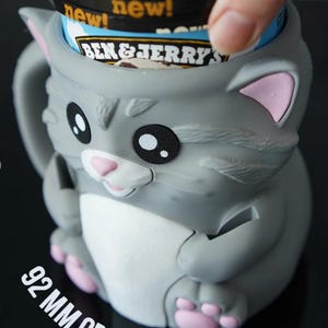 May include: A gray cat-shaped ice cream container with pink accents. The container holds a Ben & Jerry's ice cream pint. The opening is 92 mm. The container has a handle and is designed to hold a pint of ice cream.