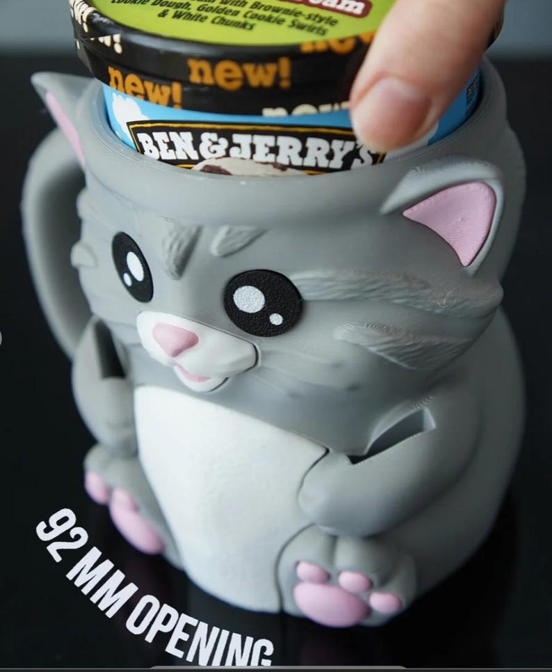 May include: A gray cat-shaped ice cream container with pink accents. The container holds a Ben & Jerry's ice cream pint. The opening is 92 mm. The container has a handle and is designed to hold a pint of ice cream.