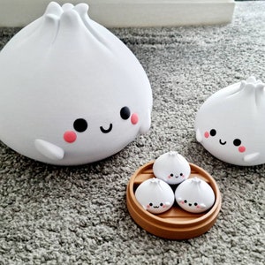 May include: Four white ceramic dumplings with smiling faces. The larger dumplings are on a white surface, while the smaller dumplings are in a brown bamboo steamer.