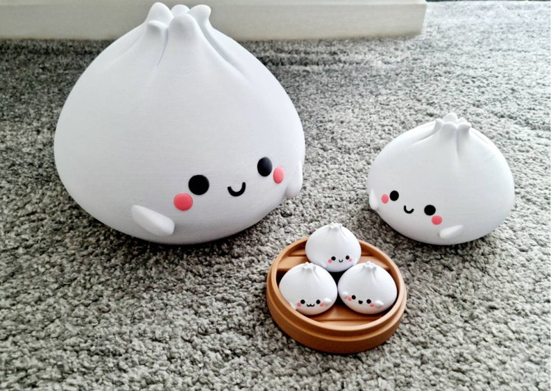 May include: Four white ceramic dumplings with smiling faces. The larger dumplings are on a white surface, while the smaller dumplings are in a brown bamboo steamer.
