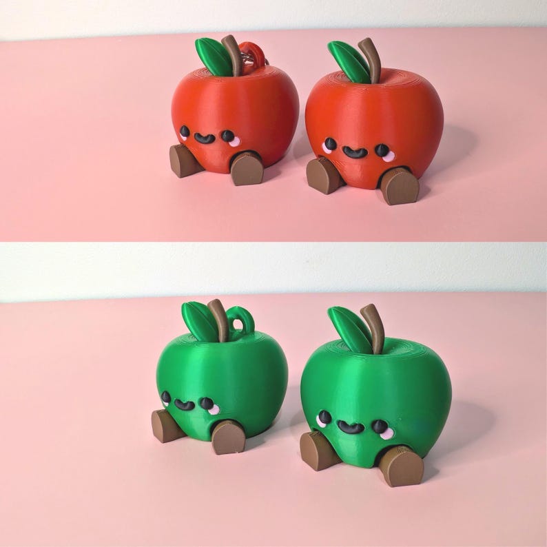 May include: Two sets of cute apple figurines, each with a smiling face, brown feet, and green leaves. One set is red, the other is green. The apples are sitting on a pink surface.