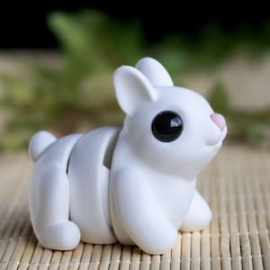 Puede incluir: Una figurita de conejo blanca segmentada con ojos negros y una nariz rosa. El conejo está sentado en una estera de bambú tejida.