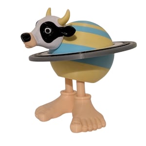 May include: A whimsical figurine featuring a cow head with a black and white face, yellow horns, and a planet-like body with blue and yellow stripes. The figurine has a gray ring around the planet and tan feet.