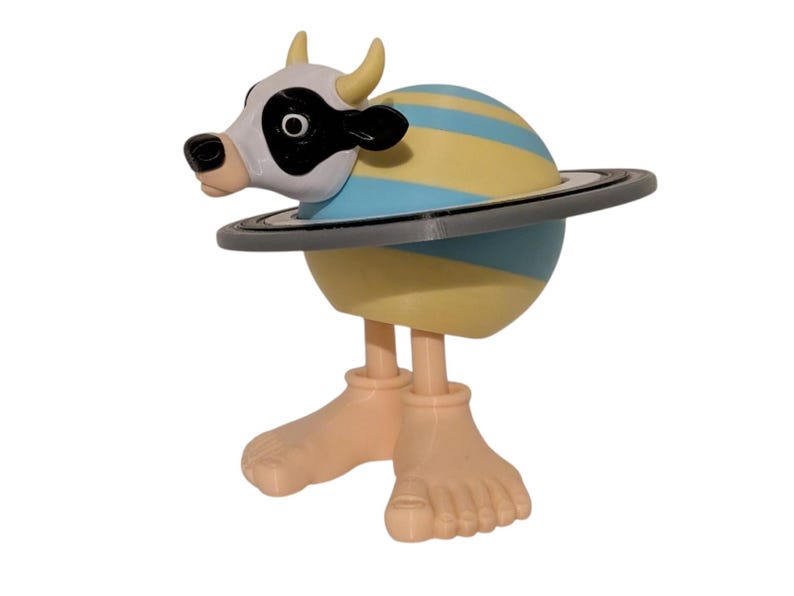 May include: A whimsical figurine featuring a cow head with a black and white face, yellow horns, and a planet-like body with blue and yellow stripes. The figurine has a gray ring around the planet and tan feet.
