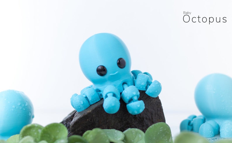 May include: A blue baby octopus toy with black eyes and a small smile. The octopus is sitting on a black rock with green leaves in the foreground. The text "Baby Octopus" is in the upper right corner of the image.