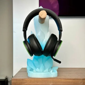 May include: A black and green gaming headset is displayed on a blue crystal-shaped stand. The stand is made of plastic and has a wood base.