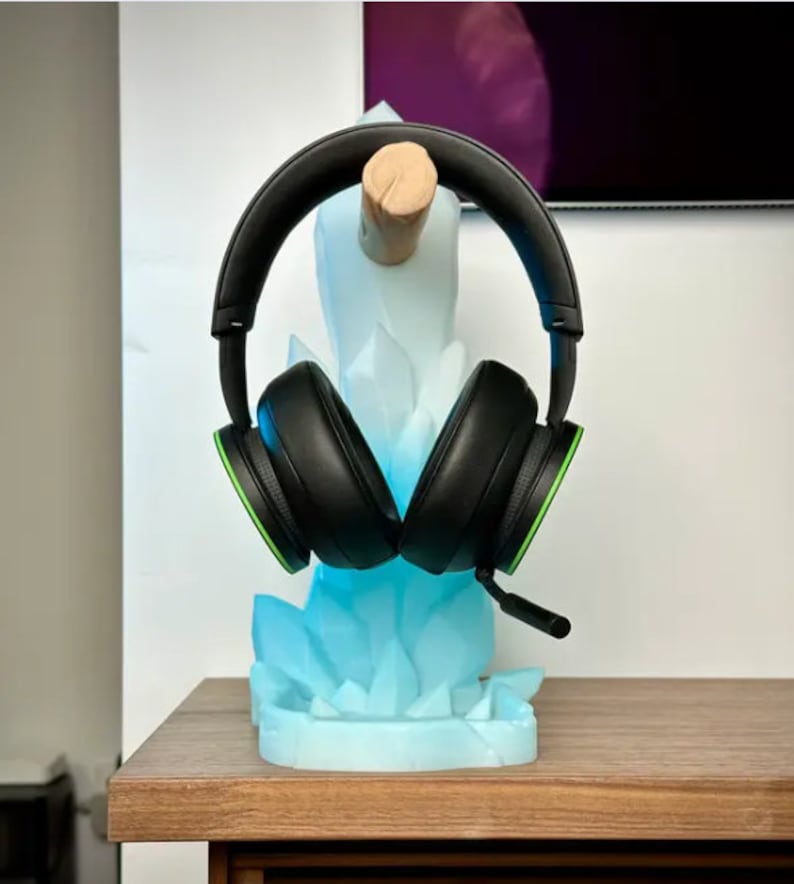 May include: A black and green gaming headset is displayed on a blue crystal-shaped stand. The stand is made of plastic and has a wood base.