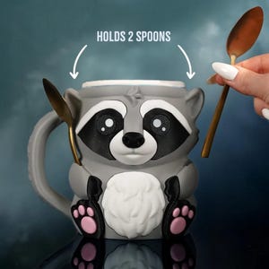 May include: Gray raccoon-shaped mug with black and white facial features, pink paw pads, and a white chest. The mug holds two gold spoons. Text above the mug reads "HOLDS 2 SPOONS".