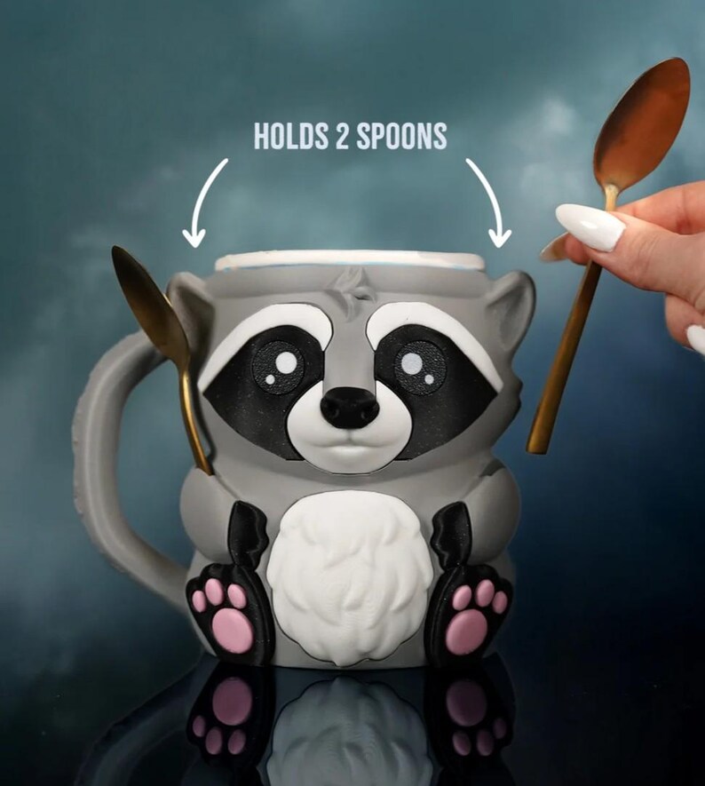 May include: Gray raccoon-shaped mug with black and white facial features, pink paw pads, and a white chest. The mug holds two gold spoons. Text above the mug reads "HOLDS 2 SPOONS".