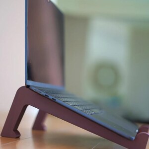 Peut inclure: Un ordinateur portable est pos&eacute; sur un support en bois de couleur bordeaux. Le support &eacute;l&egrave;ve l'ordinateur portable en angle, offrant un soutien ergonomique. L'ordinateur a un &eacute;cran et un clavier sombres.