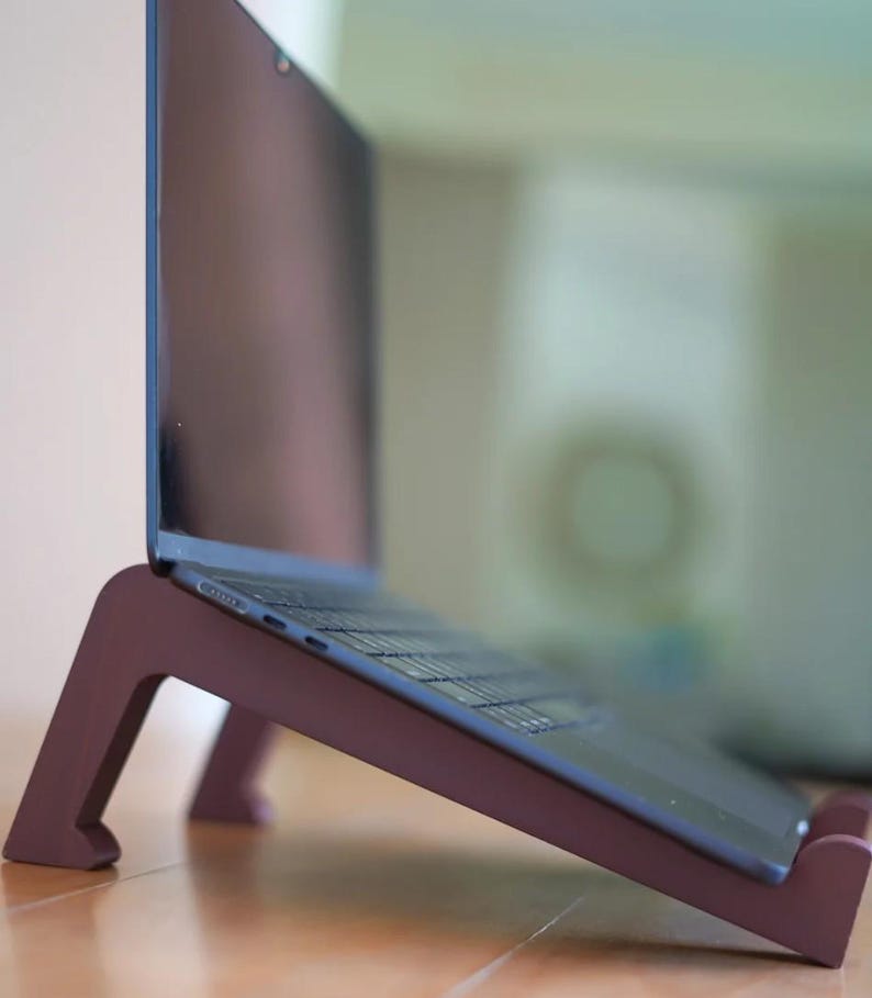 Peut inclure: Un ordinateur portable est pos&eacute; sur un support en bois de couleur bordeaux. Le support &eacute;l&egrave;ve l'ordinateur portable en angle, offrant un soutien ergonomique. L'ordinateur a un &eacute;cran et un clavier sombres.