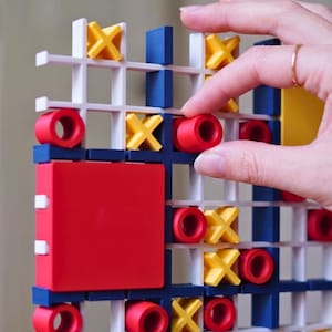 May include: A vertical tic-tac-toe game board featuring a white grid structure with blue accents. The game includes red cylindrical O pieces and yellow X pieces. A large red square block is positioned on the left side of the grid structure.
