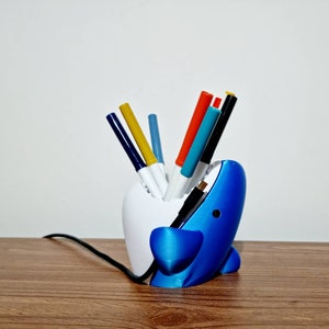 May include: A blue and white shark-shaped desk organizer with a white interior. The organizer is filled with colorful markers and pens.