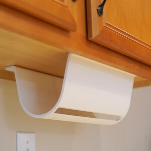 May include: A white, curved paper towel holder mounted under a wooden cabinet. The holder has a rectangular opening and is designed to dispense paper towels. The cabinet has dark handles.